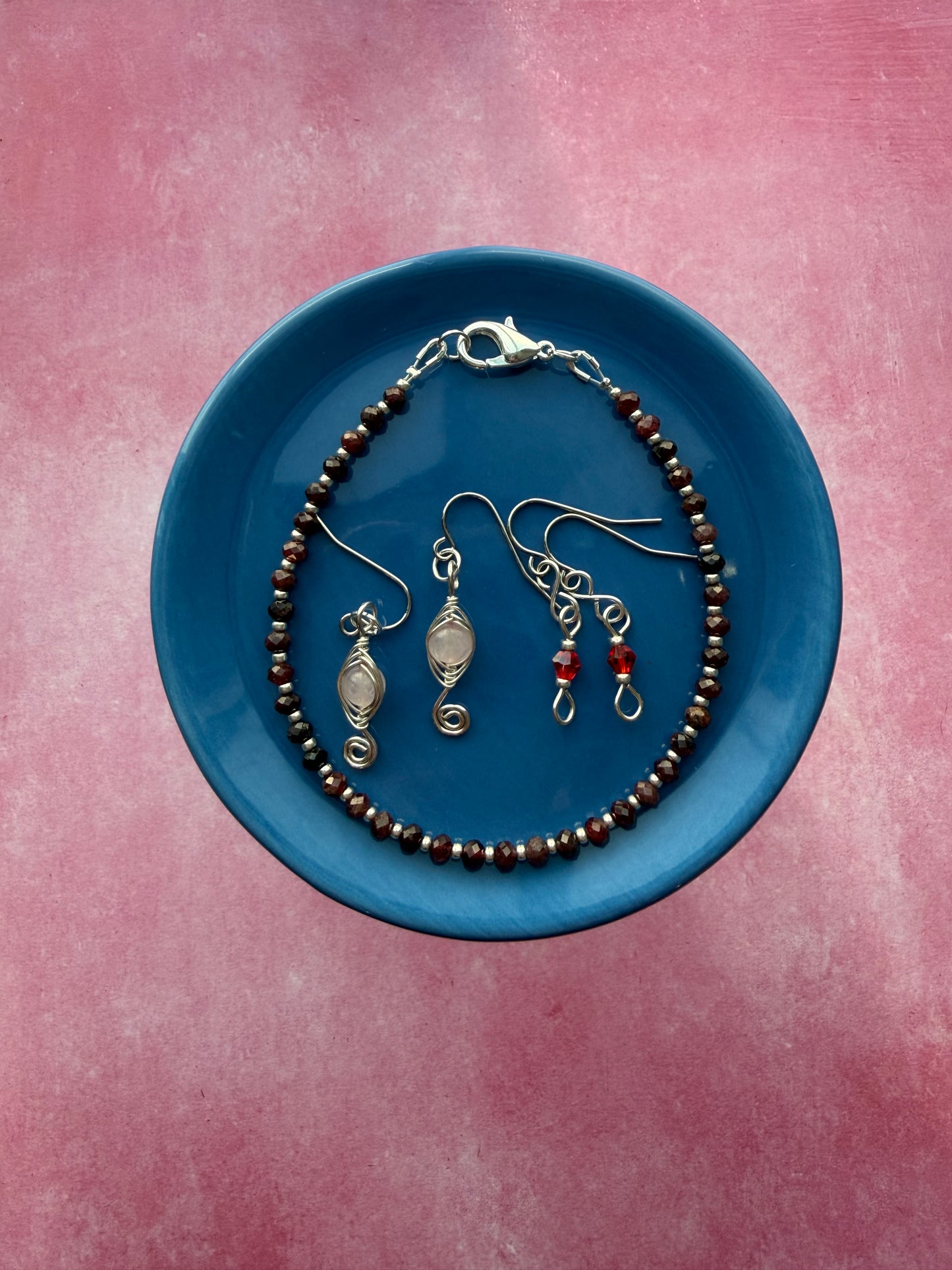 Garnet gemstone and silver seed bead stackable bracelet, red crystal petite earrings, and herringbone wire-wrapped Rose Quartz swirl earrings on a blue plate.