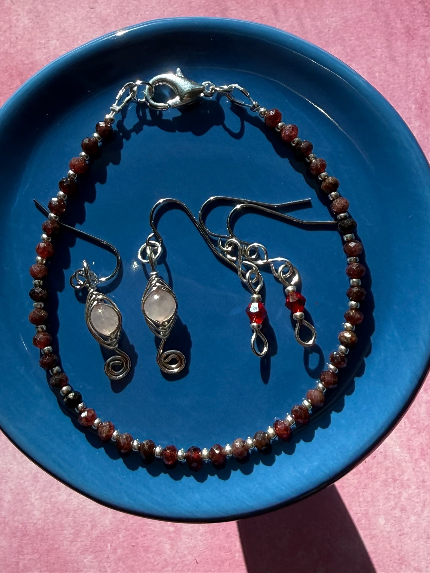 Herringbone wire-wrapped Rose Quartz swirl earrings, red crystal petite earrings, and a Garnet gemstone and silver seed bead stackable bracelet on a blue plate.