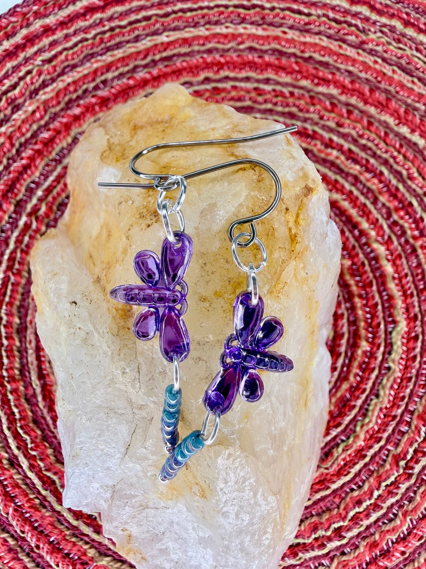 Earrings with purple dragonfly sequin charms and light blue and purple seed bead dangles lying on orange rock with red fabric background.