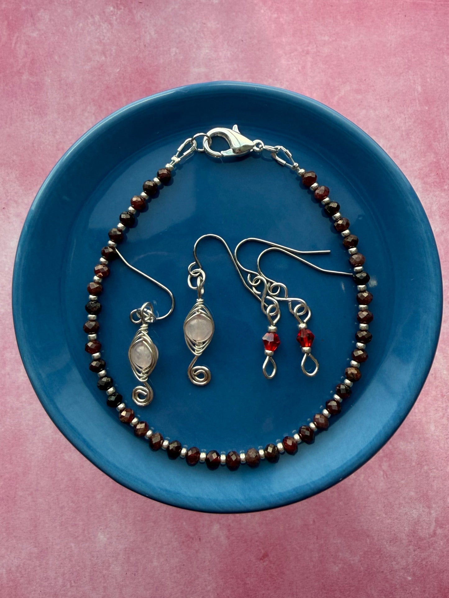 Red crystal petite earrings, herringbone wire-wrapped Rose Quartz swirl earrings, and a Garnet gemstone and silver seed bead stackable bracelet on a blue plate.