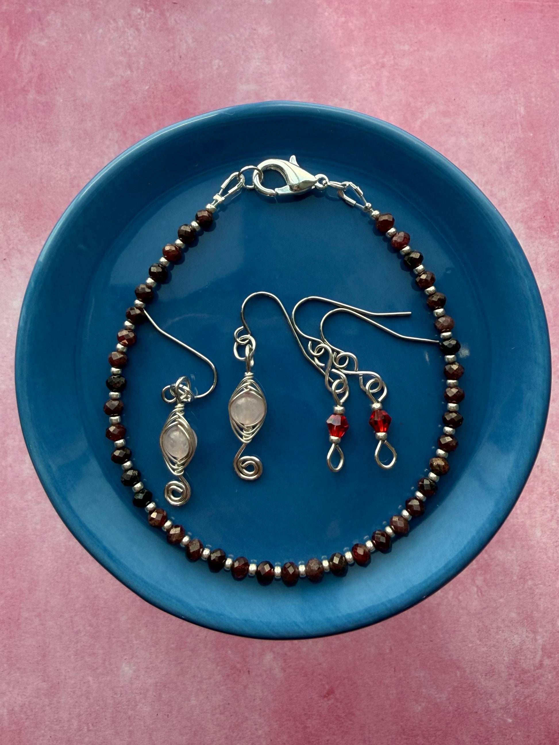 Red crystal petite earrings, herringbone wire-wrapped Rose Quartz swirl earrings, and a Garnet gemstone and silver seed bead stackable bracelet on a blue plate.
