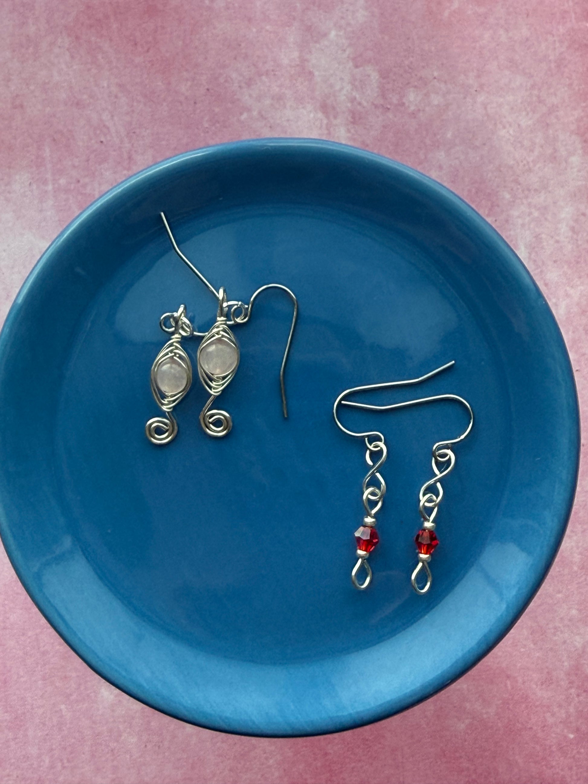 Red crystal petite earrings and herringbone wire-wrapped Rose Quartz swirl earrings on a blue plate.