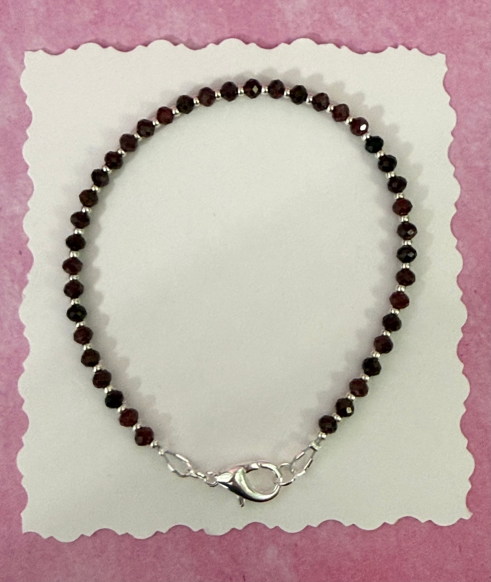 Garnet gemstone and silver seed bead stackable bracelet with a silver-plated brass lobster claw clasp on white paper with a pink background.