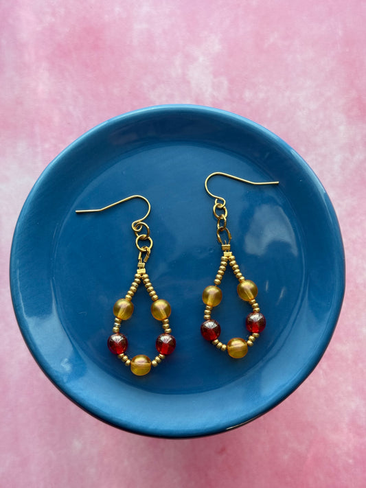 Pair of orange and red pearl beaded loop earrings with gold-plated stainless steel ear wires on a blue plate with a pink background.