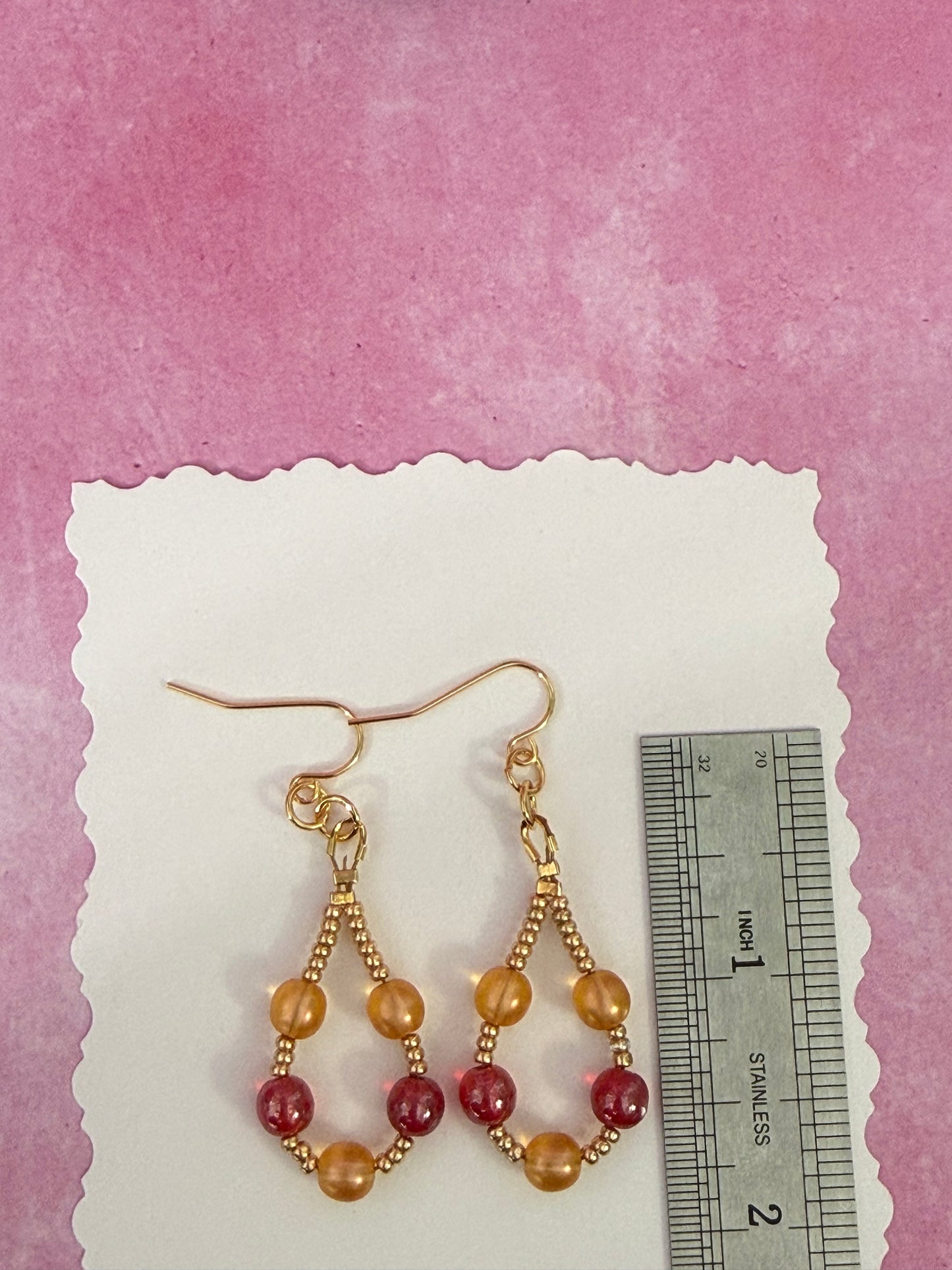 Pair of orange and red pearl beaded loop earrings with gold-plated stainless steel ear wires on a white card with a ruler for scale.
