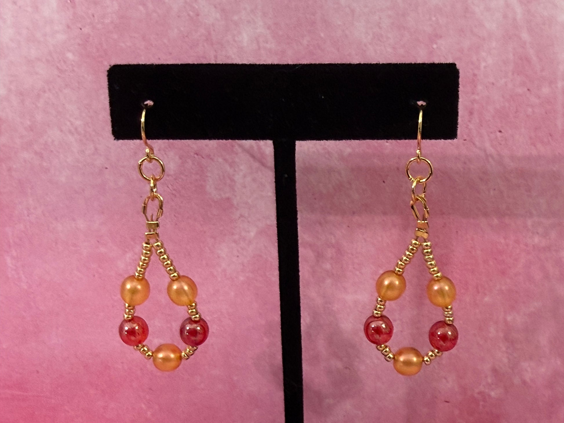 Pair of orange and red pearl beaded loop earrings with gold-plated stainless steel ear wires on a black earring stand with a pink background.