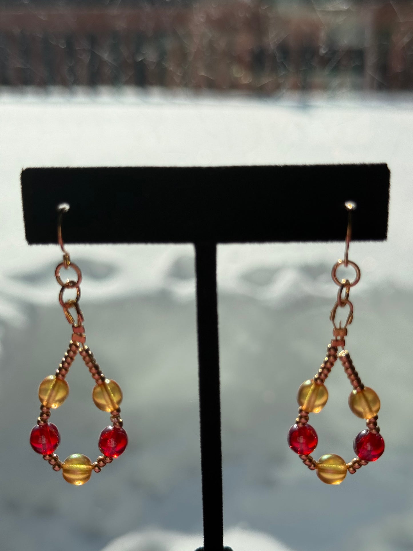 Pair of orange and red pearl beaded loop earrings with gold-plated stainless steel ear wires on a black earring stand in front of a snowy background.