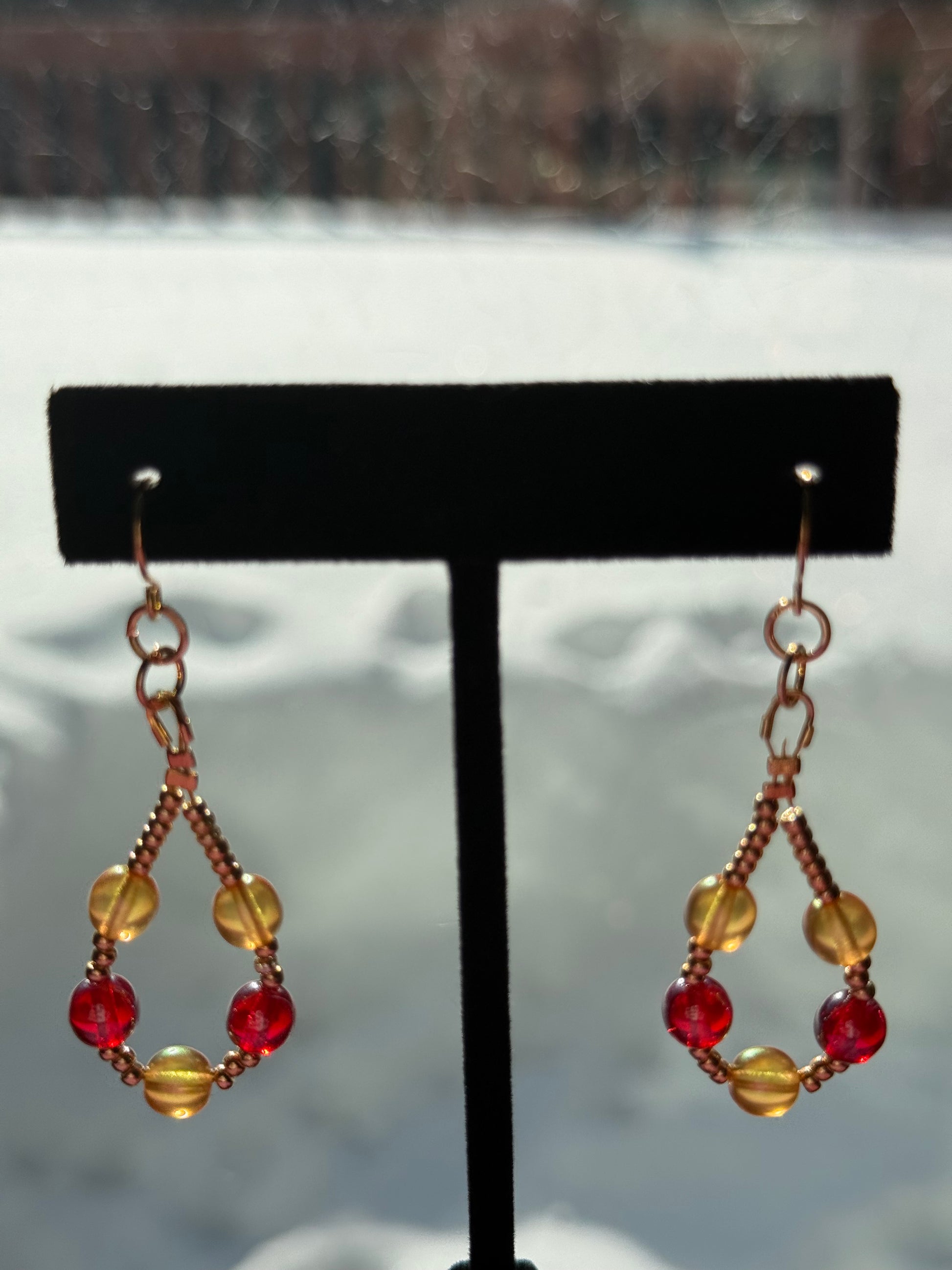 Pair of orange and red pearl beaded loop earrings with gold-plated stainless steel ear wires on a black earring stand in front of a snowy background.