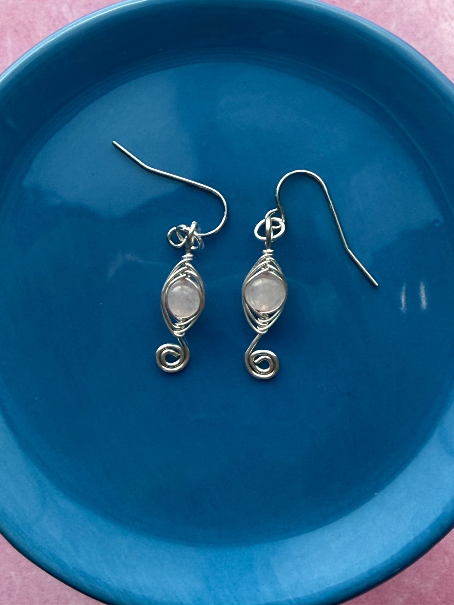 Pair of herringbone wire-wrapped earrings with rose quartz gemstone round beads and stainless steel ear wires on a blue plate with a pink background.