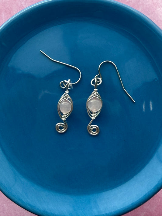 Pair of herringbone wire-wrapped earrings with rose quartz gemstone round beads and stainless steel ear wires on a blue plate with a pink background.