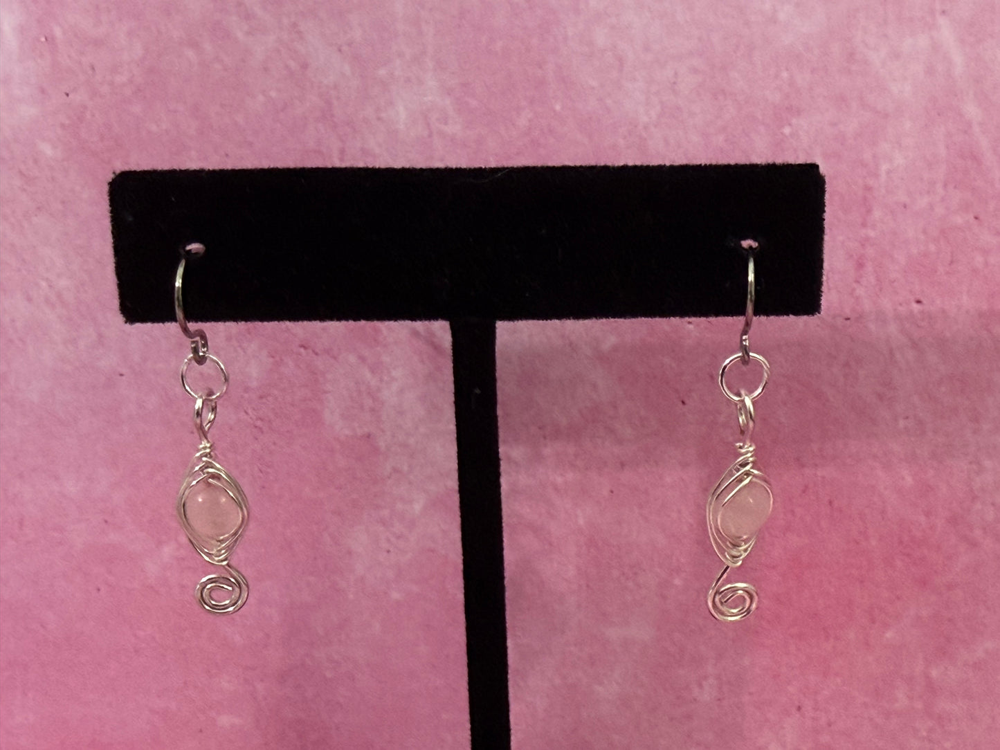Pair of herringbone wire-wrapped earrings with rose quartz gemstone round beads and stainless steel ear wires on a black earring stand with a pink background.