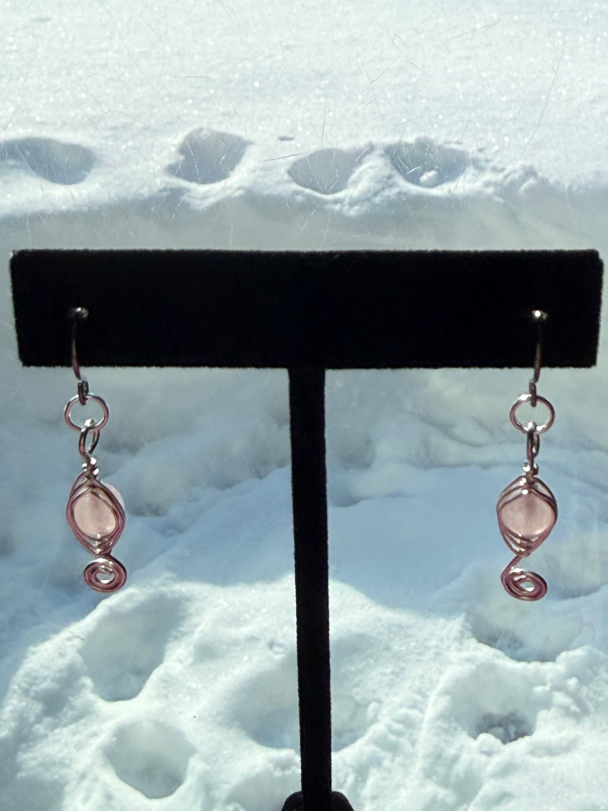Pair of herringbone wire-wrapped earrings with rose quartz gemstone round beads and stainless steel ear wires on a black earring stand in front of a snowy background.