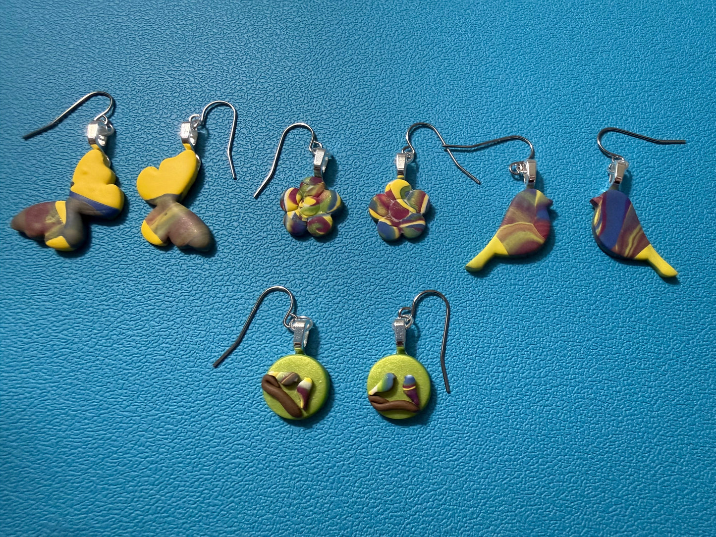 Four pairs of handcrafted polymer clay earrings from the Wildflower Meadows Sparkle and Charm Box, featuring butterfly, flower, bird, and round disc designs in swirled yellow, blue, purple, green, and white tones with stainless steel ear wires