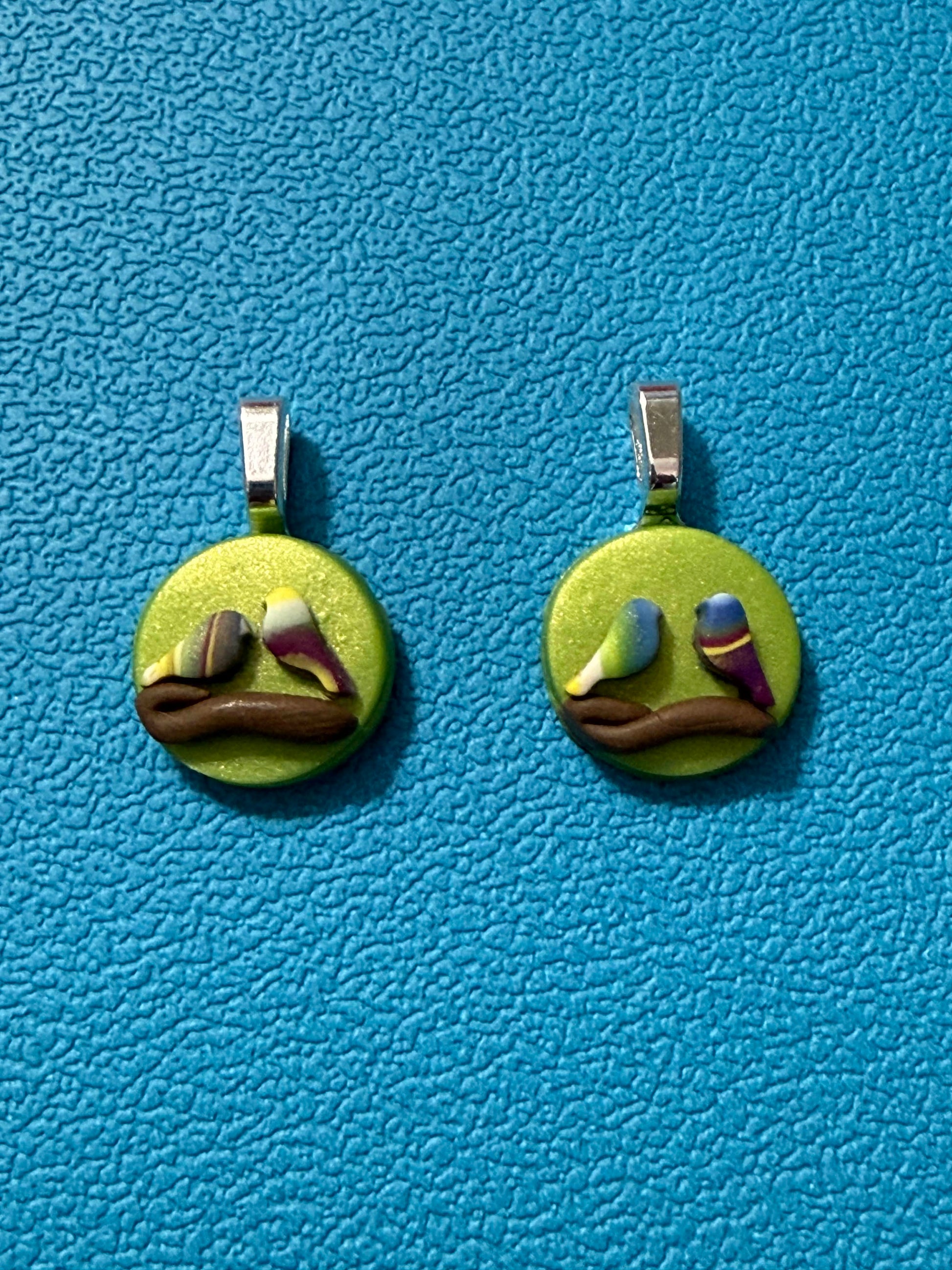 Handcrafted polymer clay round disc earrings featuring two small birds perched on a brown branch, set against a bright green background, in yellow, green, purple, blue, and white tones with stainless steel ear wires