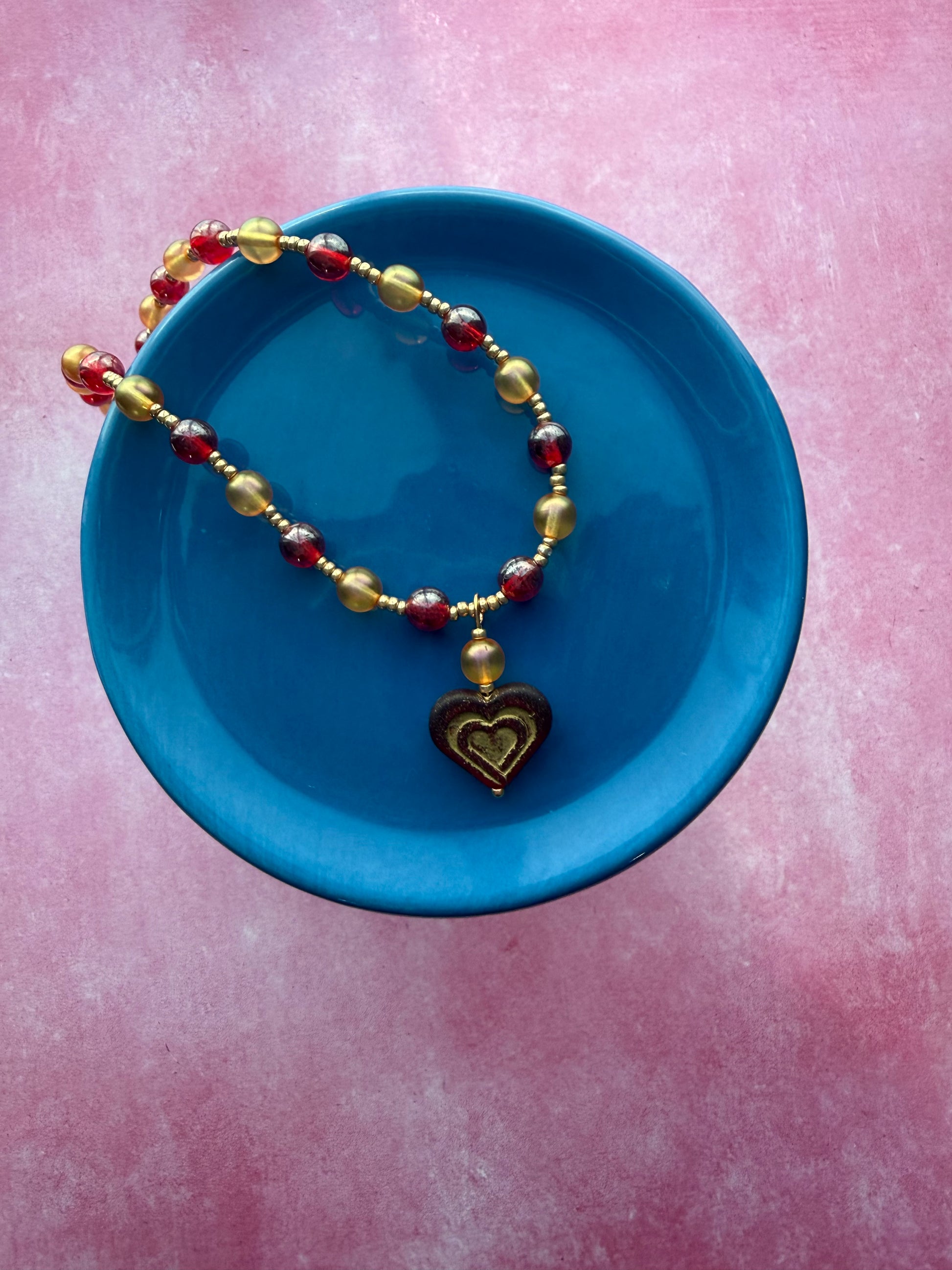 Orange and red pearl necklace with dark red and gold Czech glass heart pendant with a gold extender chain and a gold-plated brass lobster claw clasp hanging off a blue plate in a way to show off the pendant.
