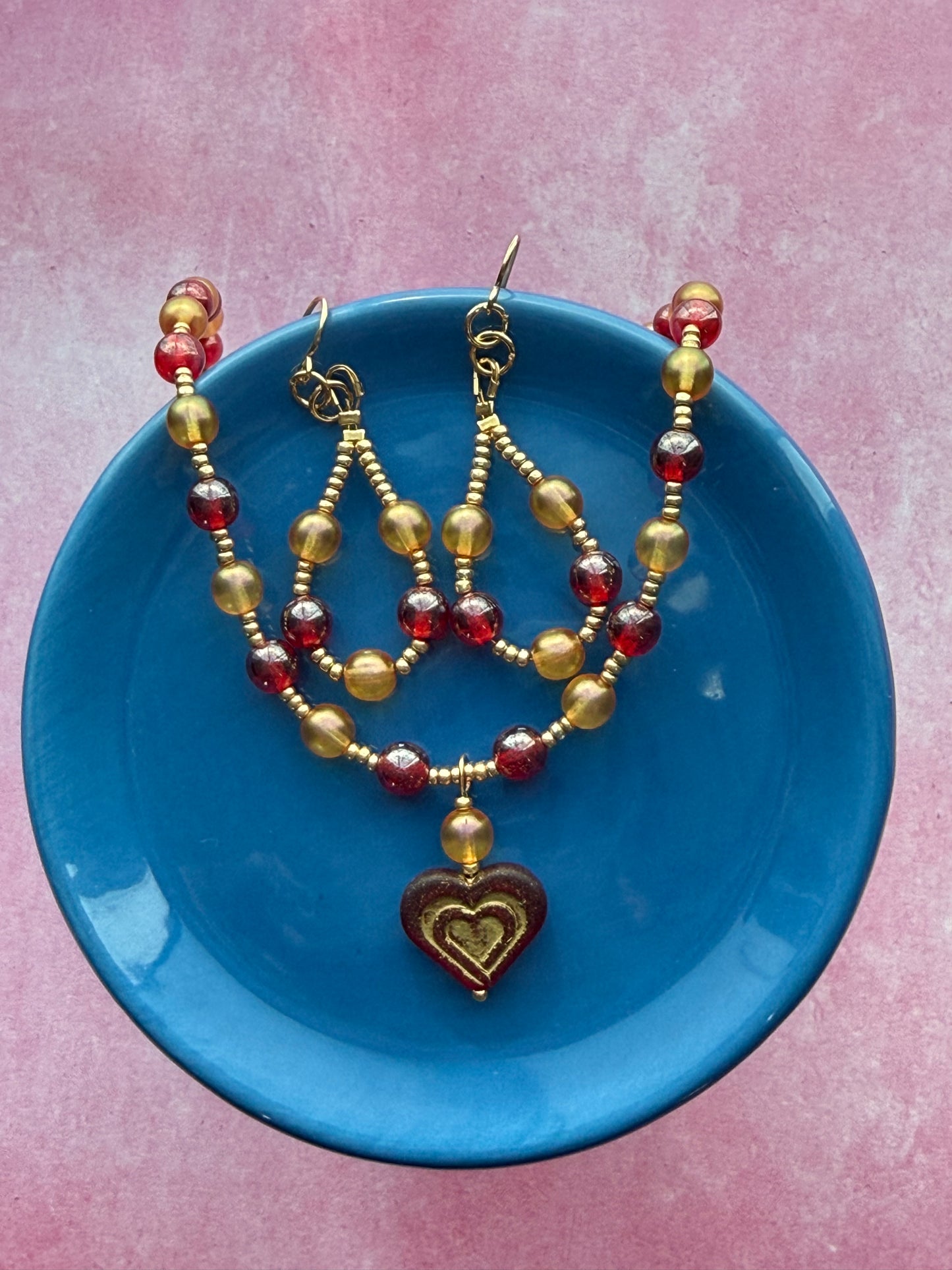 Orange and red pearl necklace with dark red and gold Czech glass heart pendant with a gold extender chain and a gold-plated brass lobster claw clasp with 0range and red pearl beaded loop earrings on a blue plate.