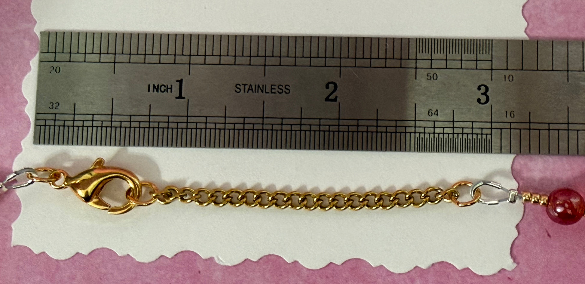 Gold extender chain of an orange and red pearl necklace on a white card with a ruler for scale.