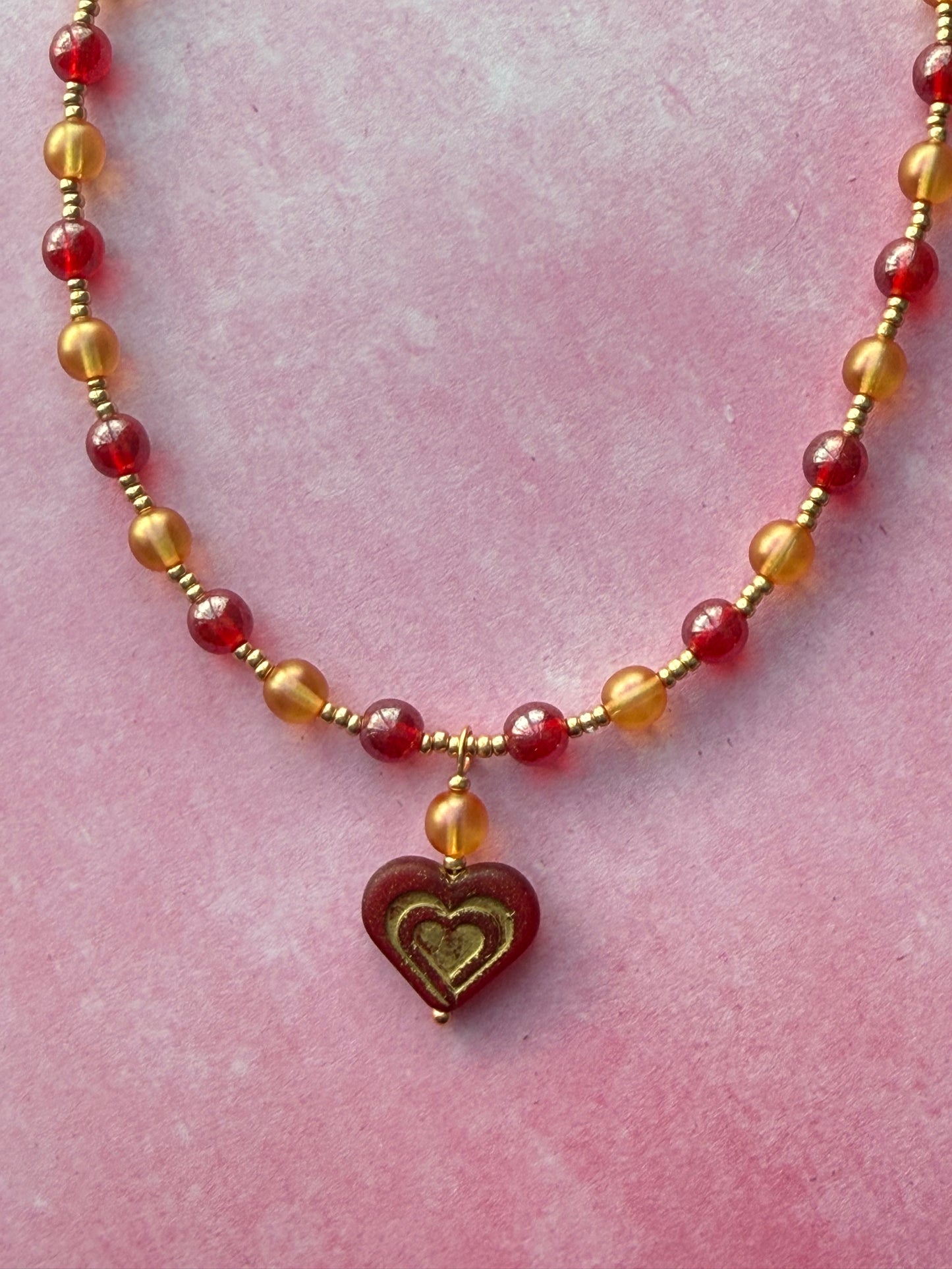 Closeup of the dark red and gold Czech glass heart pendant on a orange and red pearl necklace with a gold extender chain and gold-plated brass lobster claw clasp. Necklace is lying on a pink background.
