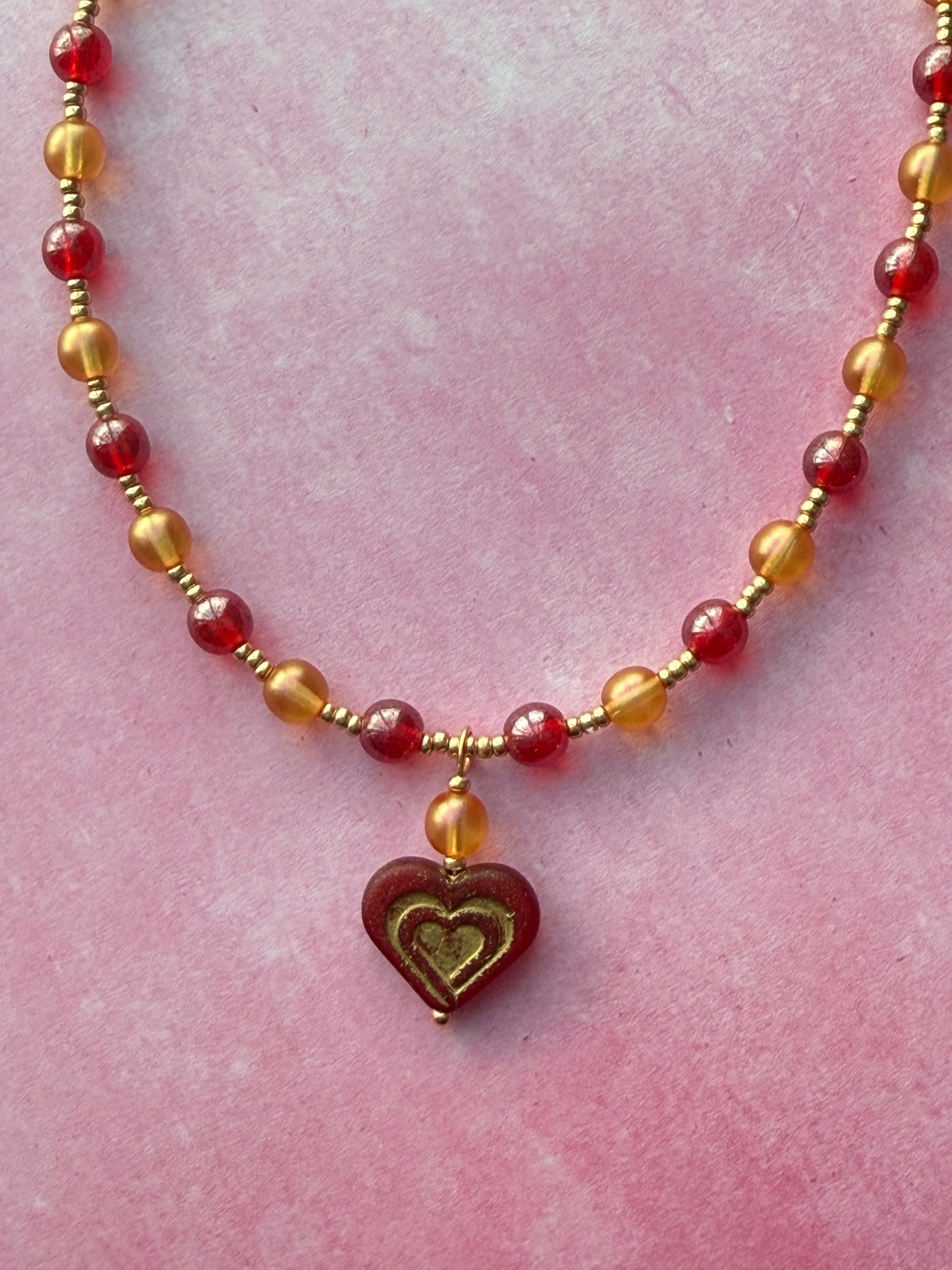 Closeup of the dark red and gold Czech glass heart pendant on a orange and red pearl necklace with a gold extender chain and gold-plated brass lobster claw clasp. Necklace is lying on a pink background.