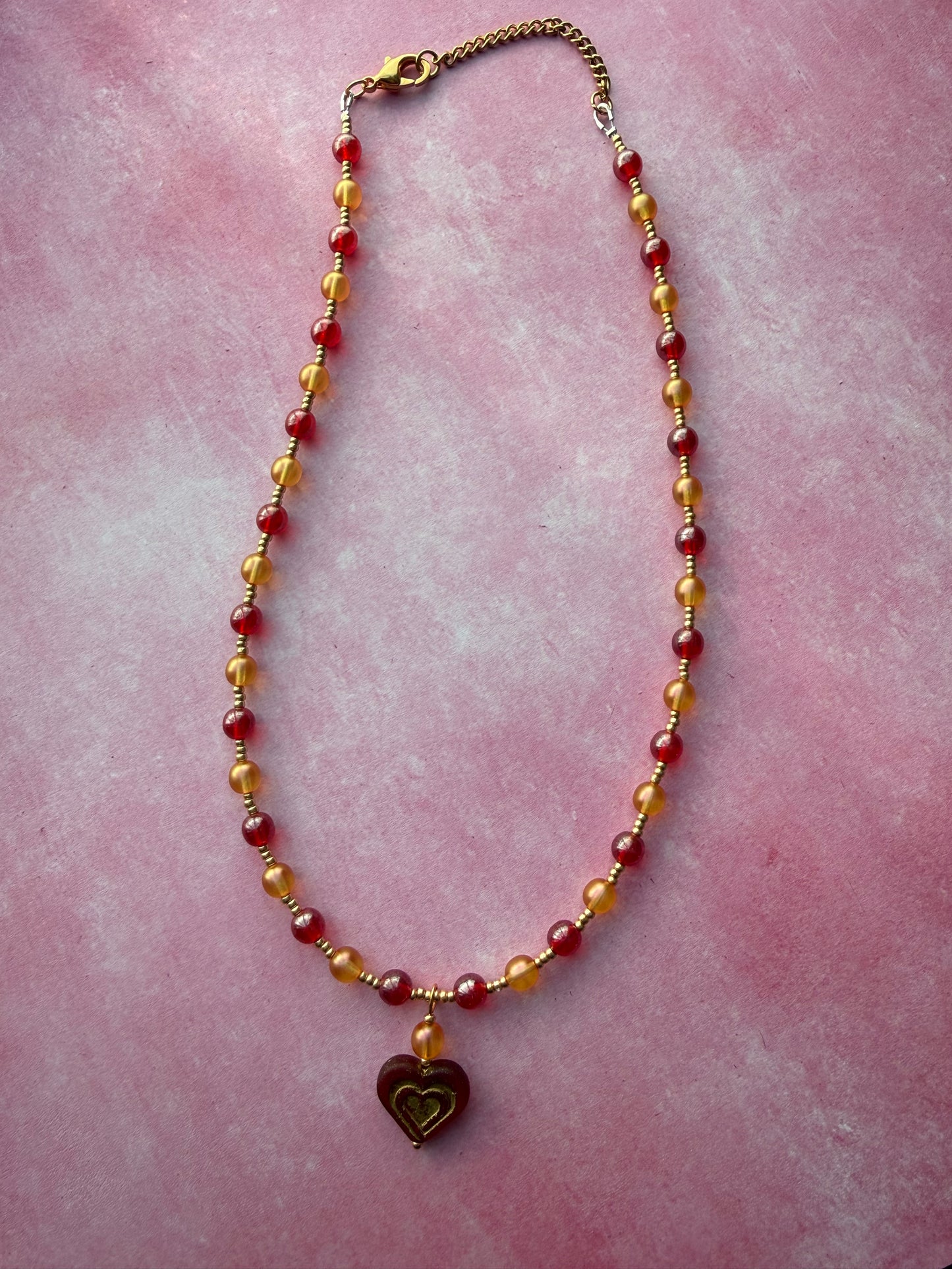 Orange and red pearl necklace with dark red and gold Czech glass heart pendant with a gold extender chain and gold-plated brass lobster claw clasp on a pink background.