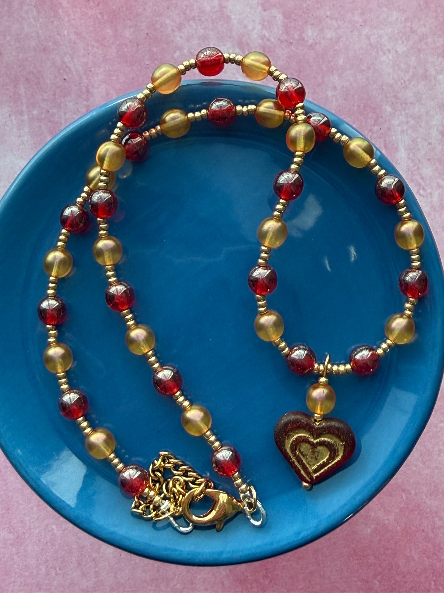 Orange and red pearl necklace with dark red and gold Czech glass heart pendant with a gold extender chain and a gold-plated brass lobster claw clasp on a blue plate.
