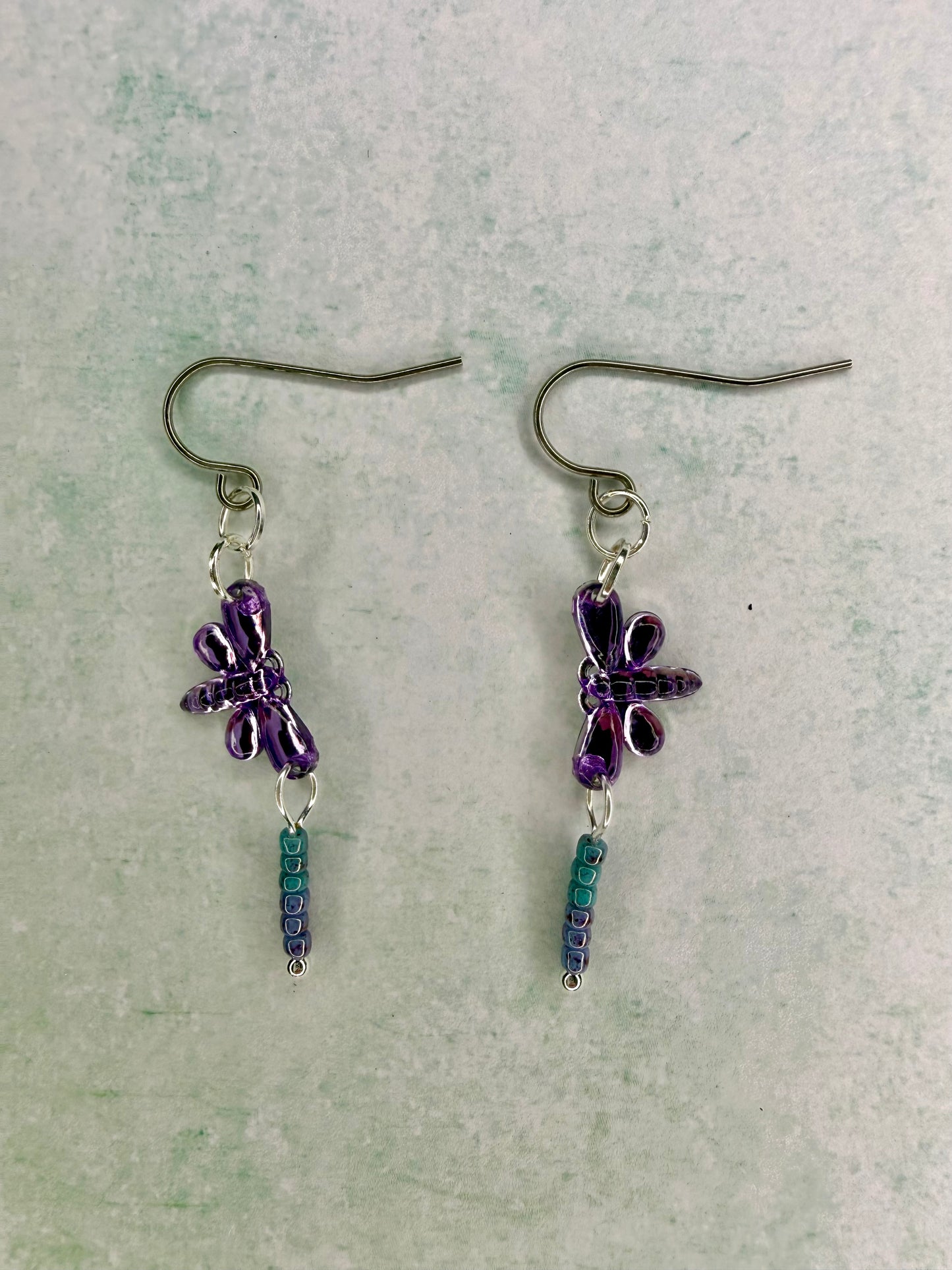 Earrings with purple dragonfly sequin charms and light blue and purple seed bead dangles lying flat on light green background.