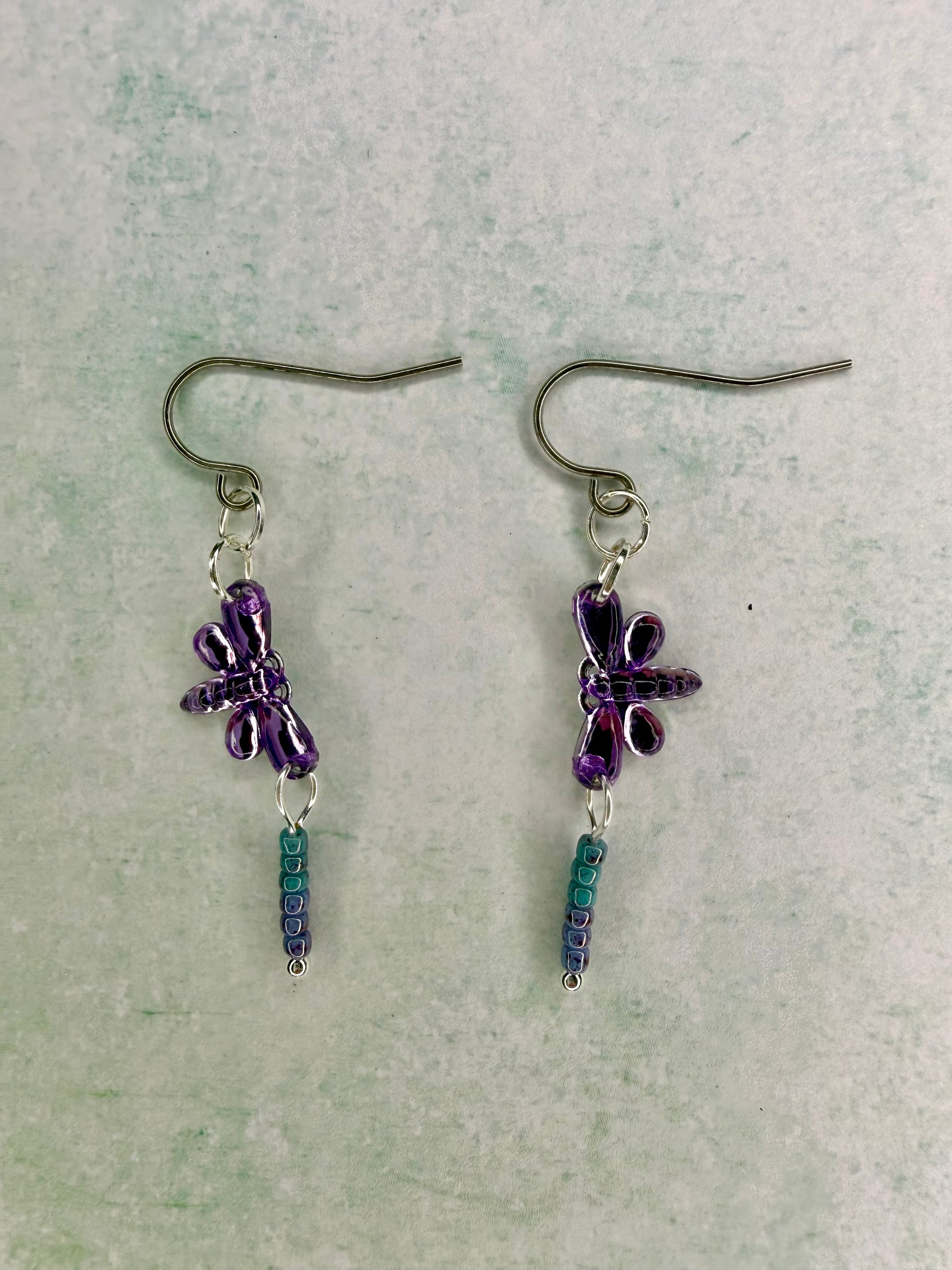 Earrings with purple dragonfly sequin charms and light blue and purple seed bead dangles lying flat on light green background.