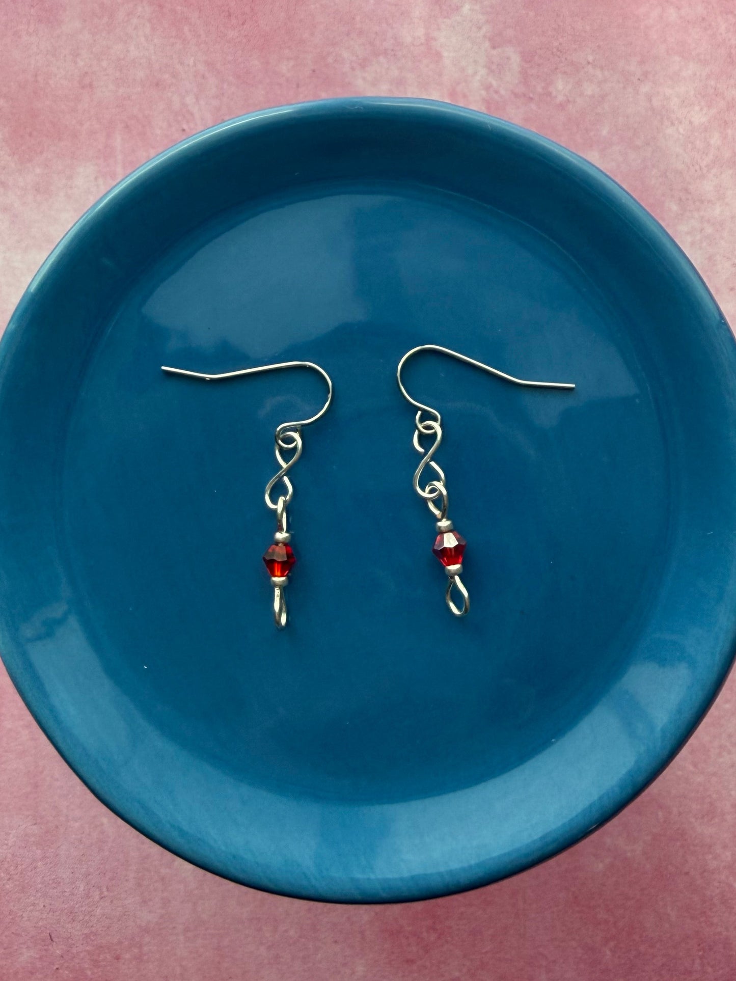 Pair of earrings with red crystal beads and stainless steel ear wires on a blue plate with a pink background.