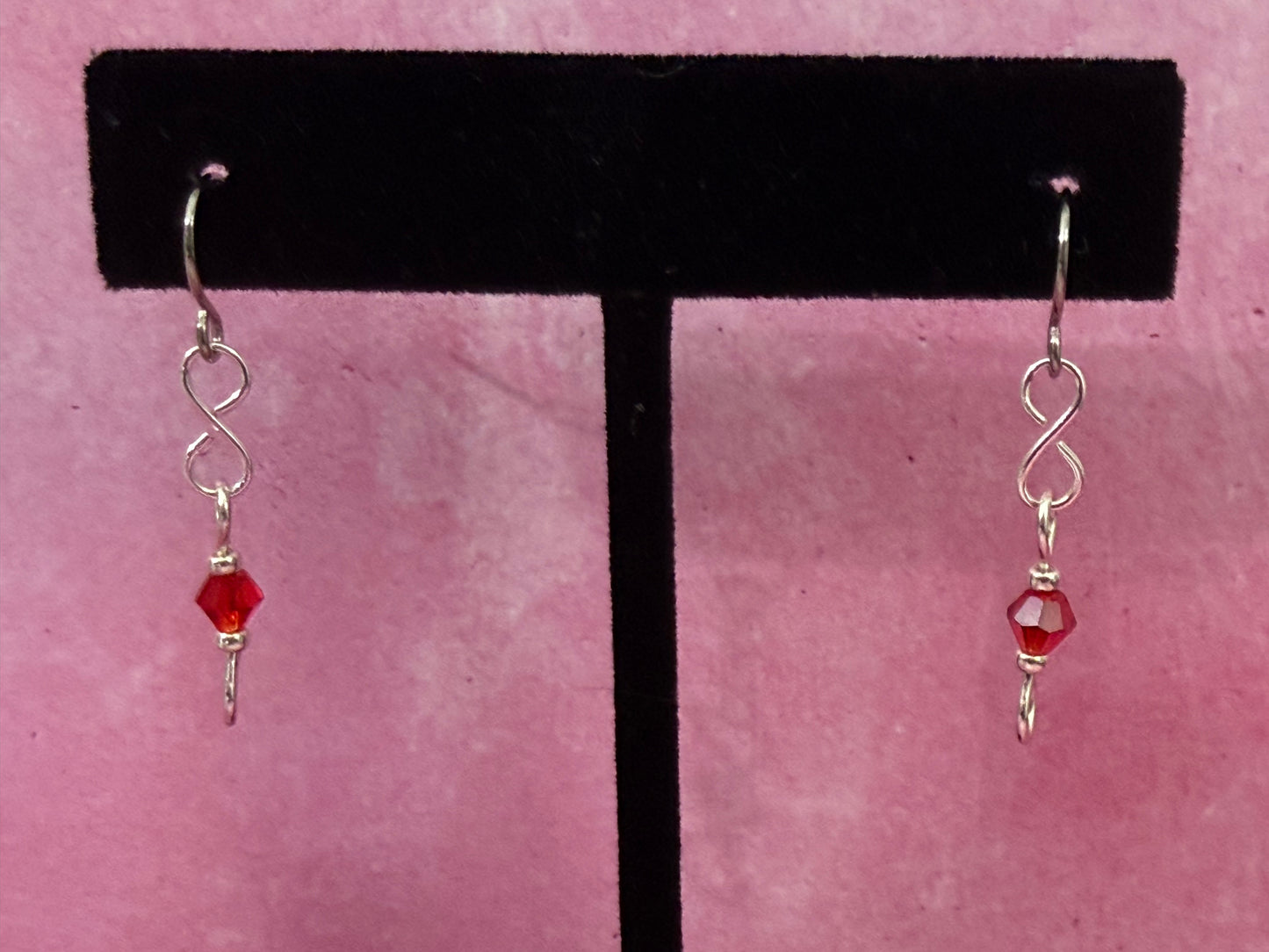 Pair of earrings with red crystal beads and stainless steel ear wires on a black earring stand with a pink background.