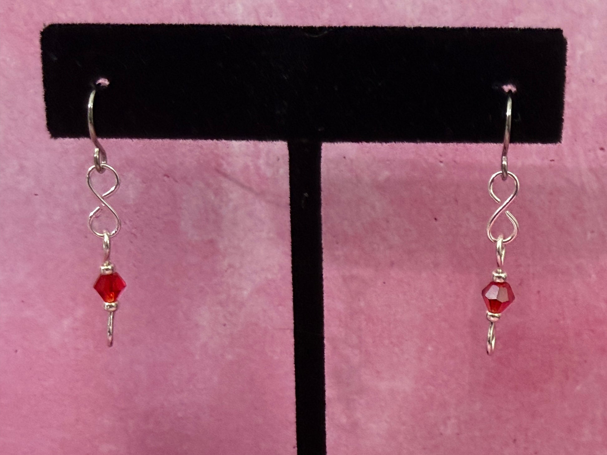 Pair of earrings with red crystal beads and stainless steel ear wires on a black earring stand with a pink background.