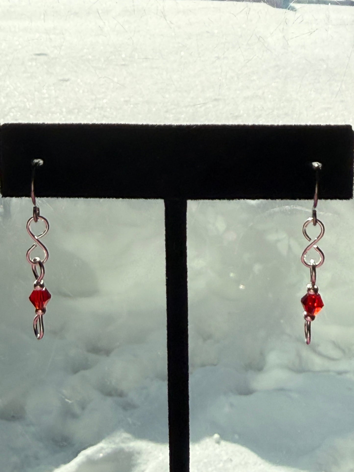 Pair of earrings with red crystal beads and stainless steel ear wires on a black earring stand in front of a snowy background.