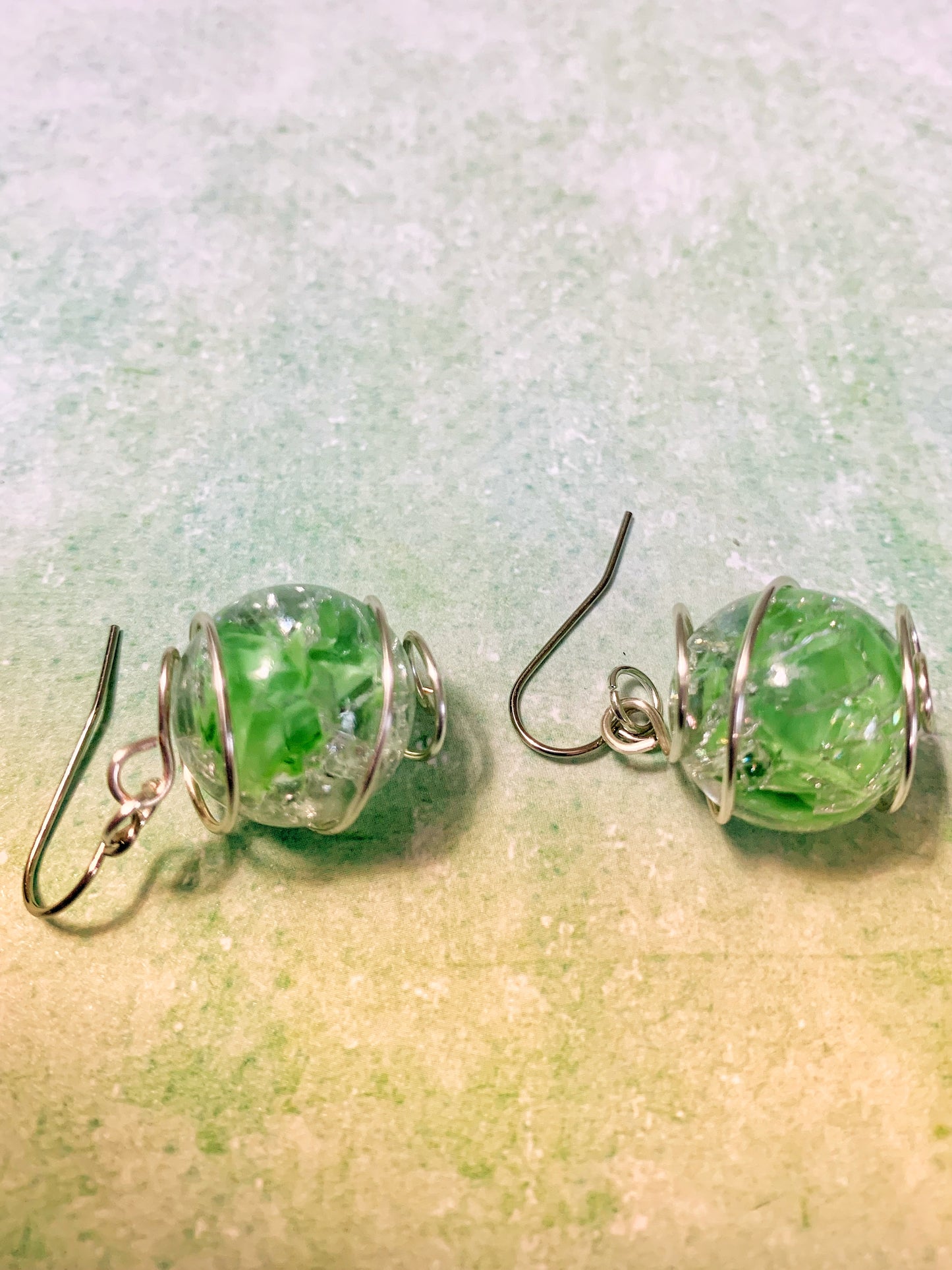 Wire-wrapped green cracked marble earrings with stainless steel ear wires lying on light green background with ear wires facing to the left.
