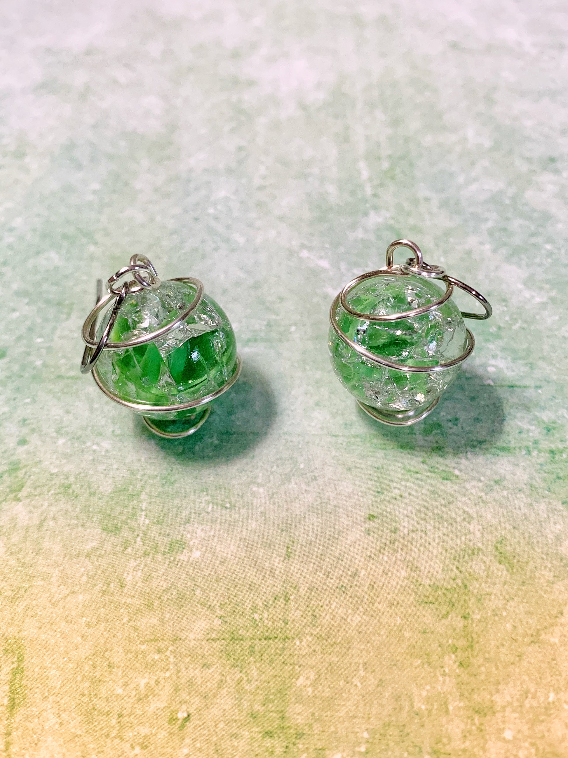 Wire-wrapped green cracked marble earrings with stainless steel ear wires lying on light green background facing up.