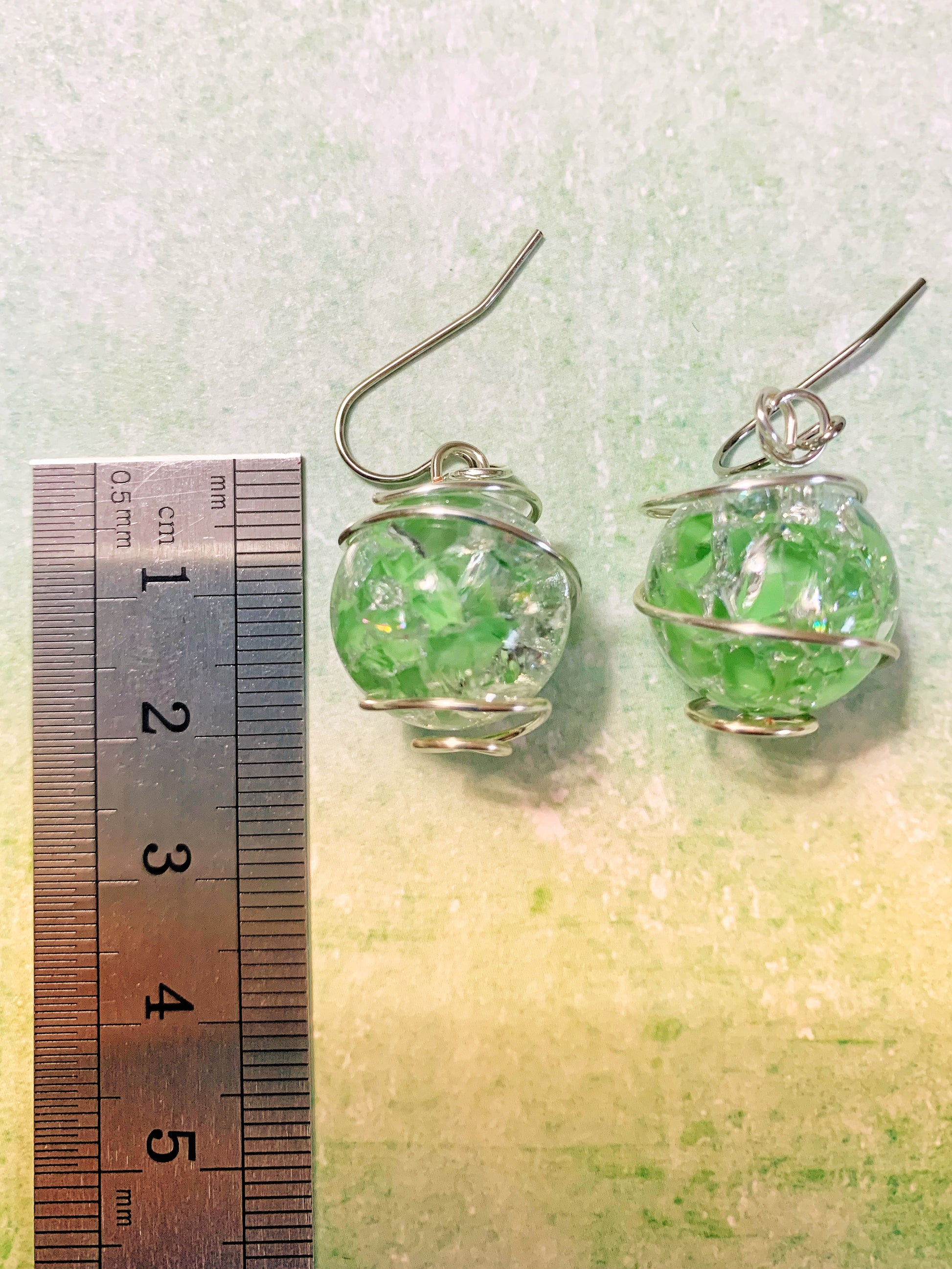Wire-wrapped green cracked marble earrings with stainless steel ear wires lying on light green background next to ruler measure about 2 inches in length.