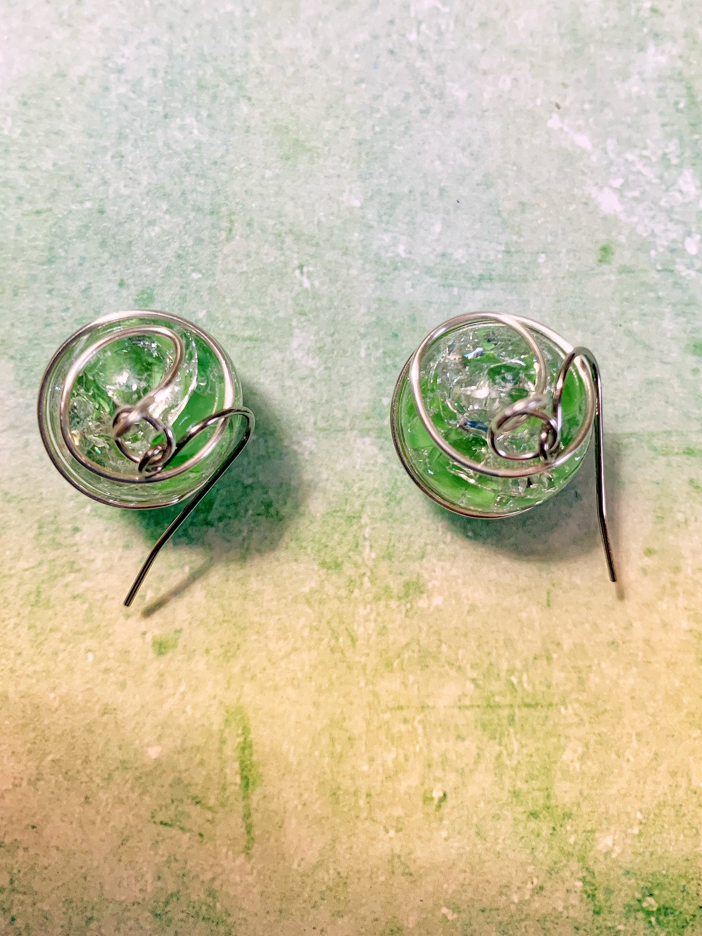 Wire-wrapped green cracked marble earrings with stainless steel ear wires lying on light green background looking from the top down.