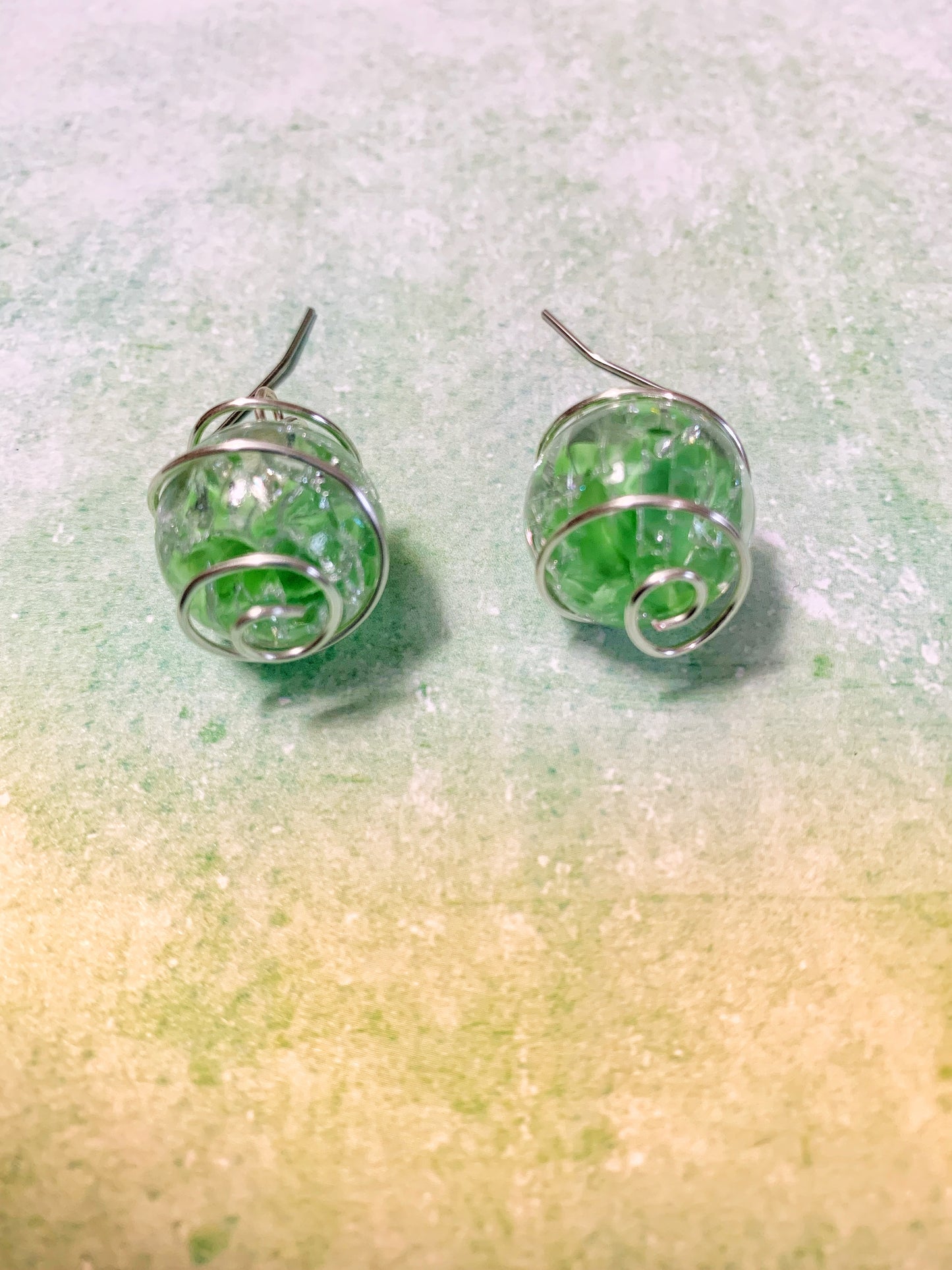 Wire-wrapped green cracked marble earrings with stainless steel ear wires lying flat on light green background with close-ups of bottom swirls.
